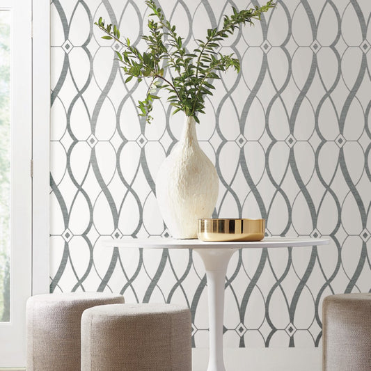 A modern, minimalist room features a white round table with a tall textured vase of green branches and a gold bowl. The wall displays York Wallcoverings' White & Silver Graceful Geo Wallpaper. Two beige stools highlight the luminous metallic finish, enhancing the elegant ambiance.