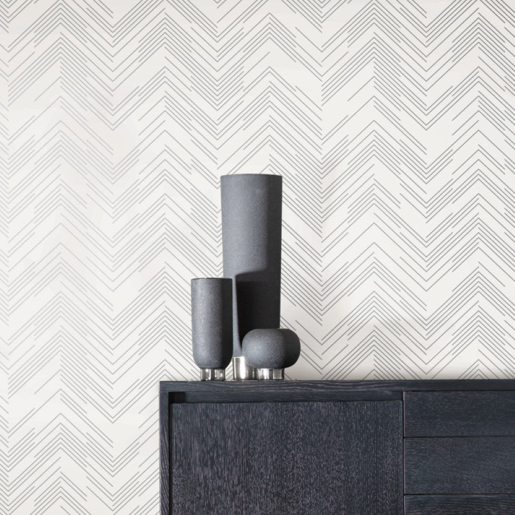 A modern interior includes a black wooden cabinet adorned with three matte black vases of varying heights. The walls feature York Wallcoverings' White & Silver Polished Chevron Wallpaper, adding textured sophistication to the minimalist decor.