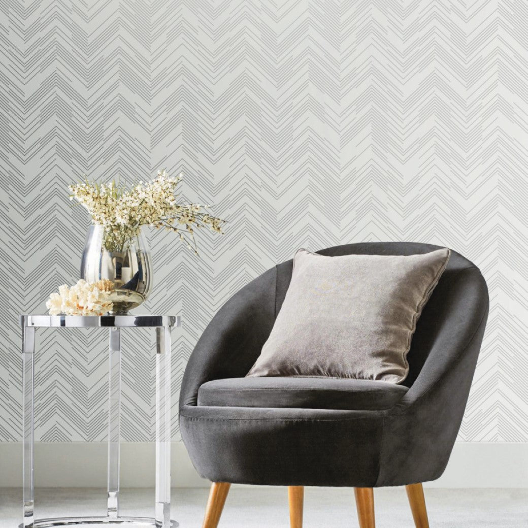 A modern room includes a gray curved chair with a matching cushion on light wooden legs. A silver side table holds a vase of white flowers, set against a white wall featuring York Wallcoverings' White & Silver Polished Chevron Wallpaper in light gray hues.