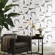 Living Room - A modern features York Wallcoverings' white & silver abstract geo wallpaper. A green armchair with a gray blanket pairs with a black side table adorned with a lamp and flowers. A large potted plant in the corner enhances the contemporary interior