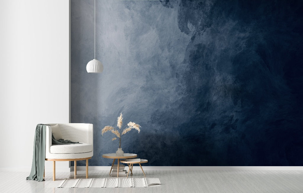 Living Room - A modern living room corner with a white armchair, a wooden side table with books and a vase with dried flowers, against a textured deep blue feature wall. A simple white pendant lamp hangs above the Decor2Go Wallpaper Mural.