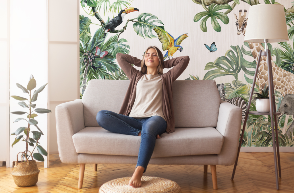 A woman relaxes on a sofa with her hands behind her head in a bright living room decorated with a Decor2Go Wallpaper Mural featuring the Wild Animals and the Jungle Watercolor design of birds and plants.