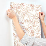 A person wearing a striped shirt is applying York Wallcoverings Willow Grove Clay Wallpaper Pink (60 Sq.Ft.) with a leafy, pastel-colored botanical design to a white wall. The SureStrip wallpaper features an elegant pattern of light pink and beige leaves, creating a serene botanical retreat.