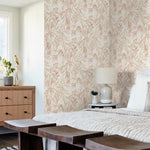 A cozy bedroom features a bed with light-colored bedding, a bedside table with a lamp, and a dresser holding a plant and decorative items. The room is adorned with York Wallcoverings' Willow Grove Clay Wallpaper Pink (60 Sq.Ft.), creating a botanical retreat as natural light streams through the window.