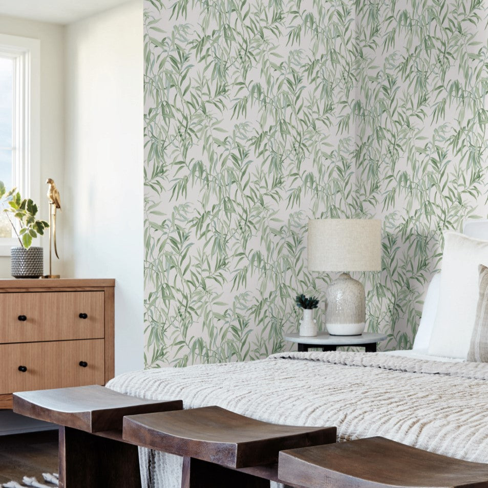 A cozy bedroom with a bed dressed in neutral bedding and wooden end benches. The back wall features an elegant York Wallcoverings Willow Grove Clay Wallpaper Pink (60 Sq.Ft.) with green and white leafy designs, transforming the space into a botanical retreat. To the left, a light wooden dresser holds a decorative lamp and potted plant near a large window allowing natural light in.