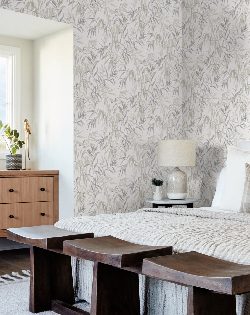 A serene bedroom featuring a bed with a knitted throw blanket and neutral tones. A wooden bench sits at the foot of the bed, while a wooden dresser with a potted plant is near the window. The walls are adorned with York Wallcoverings' Willow Grove Clay Wallpaper Beige(60 Sq.Ft.), showcasing a subtle willow trees pattern.