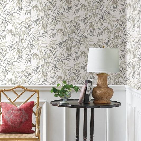 A corner of a room is shown with a round wooden side table holding a wicker table lamp with a white shade, a stack of books, and a small potted plant. The wall is adorned with York Wallcoverings Willow Grove Sand Wallpaper Beige, Grey (60 Sq.Ft.) featuring leafy patterns in muted tones, adding natural elegance. A woven chair with a red cushion completes the cozy scene.