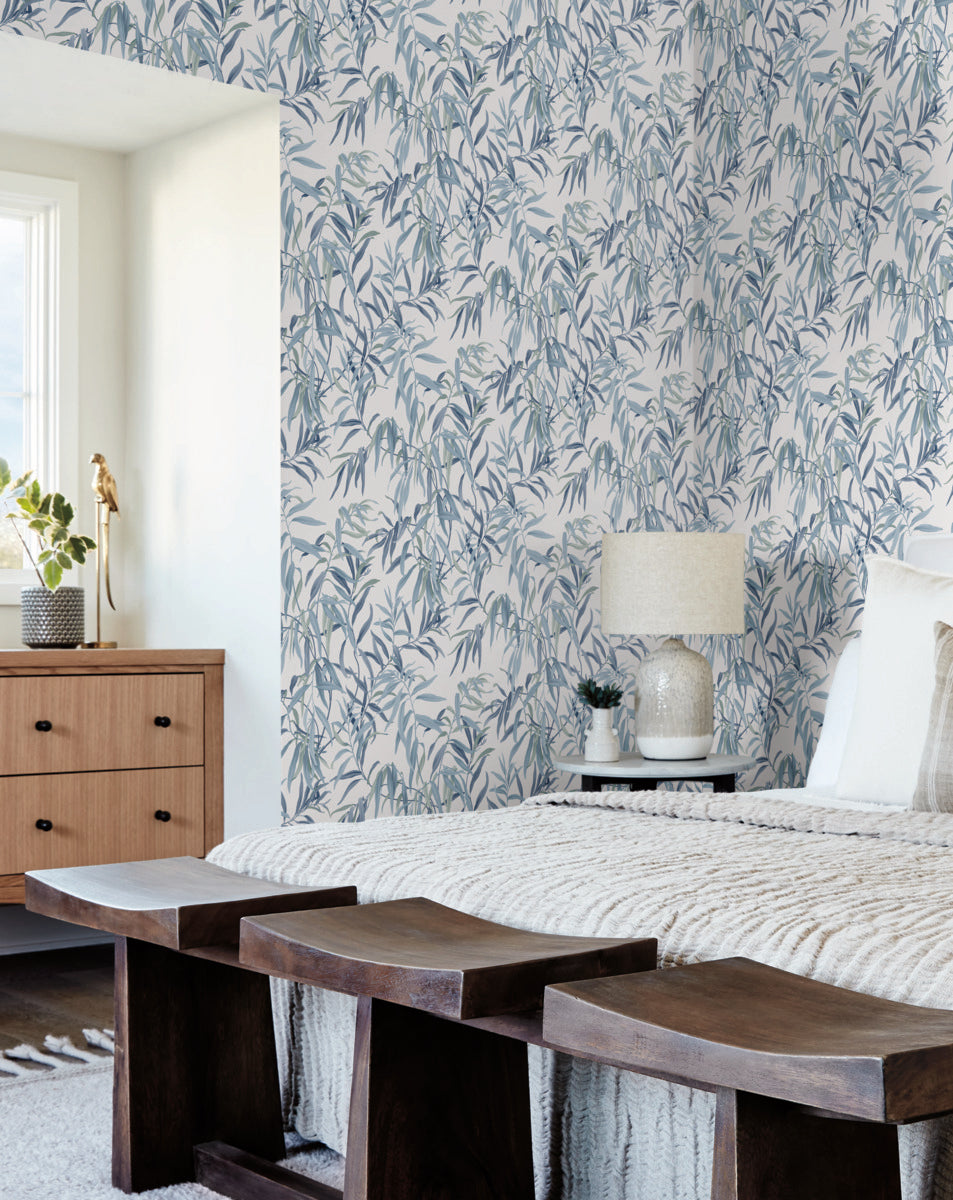 A cozy bedroom featuring a bed with a white knitted throw blanket and several pillows, a wooden bench at the foot, and a wooden dresser with a plant. The room is bathed in natural light from the window, and the walls are adorned with York Wallcoverings Willow Grove Clay Wallpaper Pink (60 Sq.Ft.), creating a botanical retreat. A table lamp sits on the dresser.