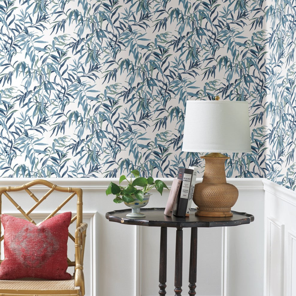 A decorative corner featuring a round, dark wooden side table with books, a potted plant, and a wicker lamp with a white shade. A bamboo chair with a red pillow sits beside the table. The background, adorned with York Wallcoverings Willow Grove Clay Wallpaper Pink (60 Sq.Ft.), completes this botanical retreat.