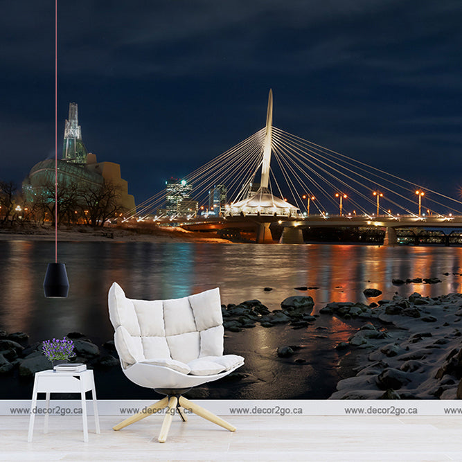 A stylized room with a white modern chair and a small table overlooking a romantic night landscape of a cityscape with illuminated Provencher Bridge and buildings reflected in the river featuring the Decor2Go Wallpaper Mural "Winnipeg in Blues.