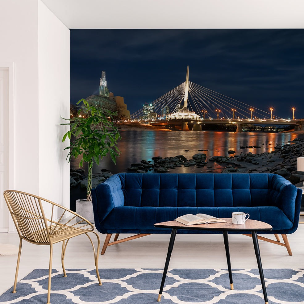 A stylish living room featuring a blue velvet sofa, a black circular table with a book and cup, and a gold chair, facing a large window showcasing a night cityscape with the lit-up Decor2Go Wallpaper Mural.