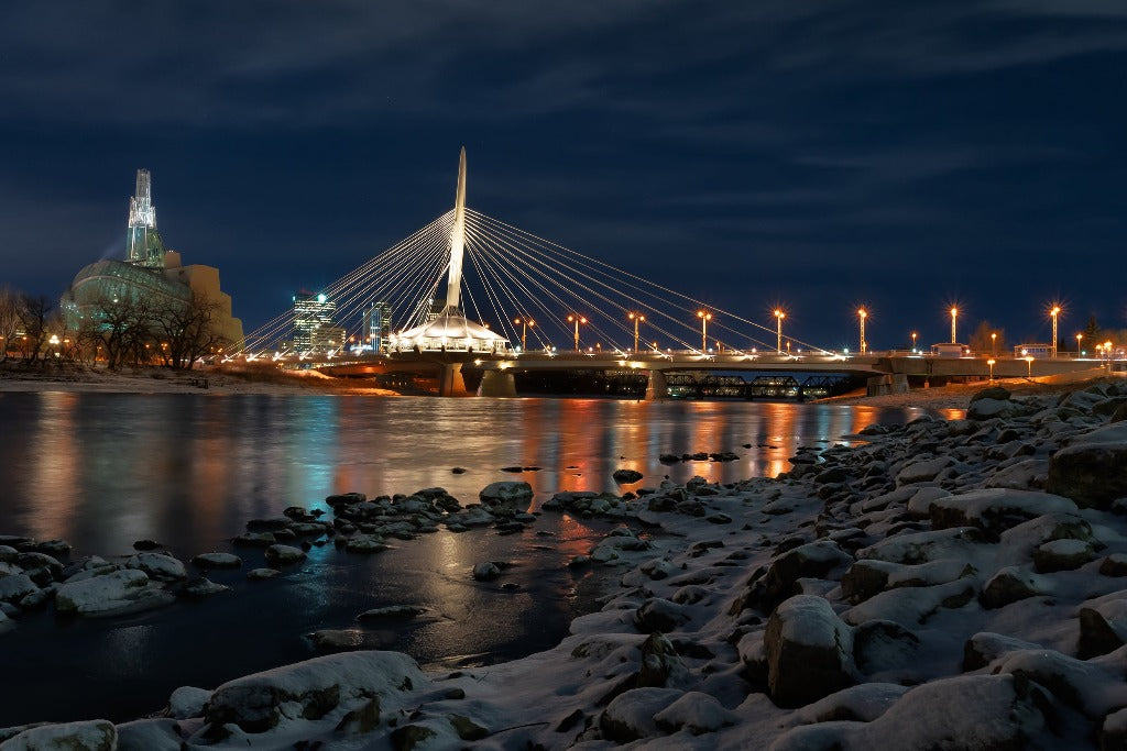 A nighttime view of the modern Decor2Go Wallpaper Mural illuminated over a partially frozen river, with city buildings lit up in the background and snowy, rocky riverbanks in the foreground.