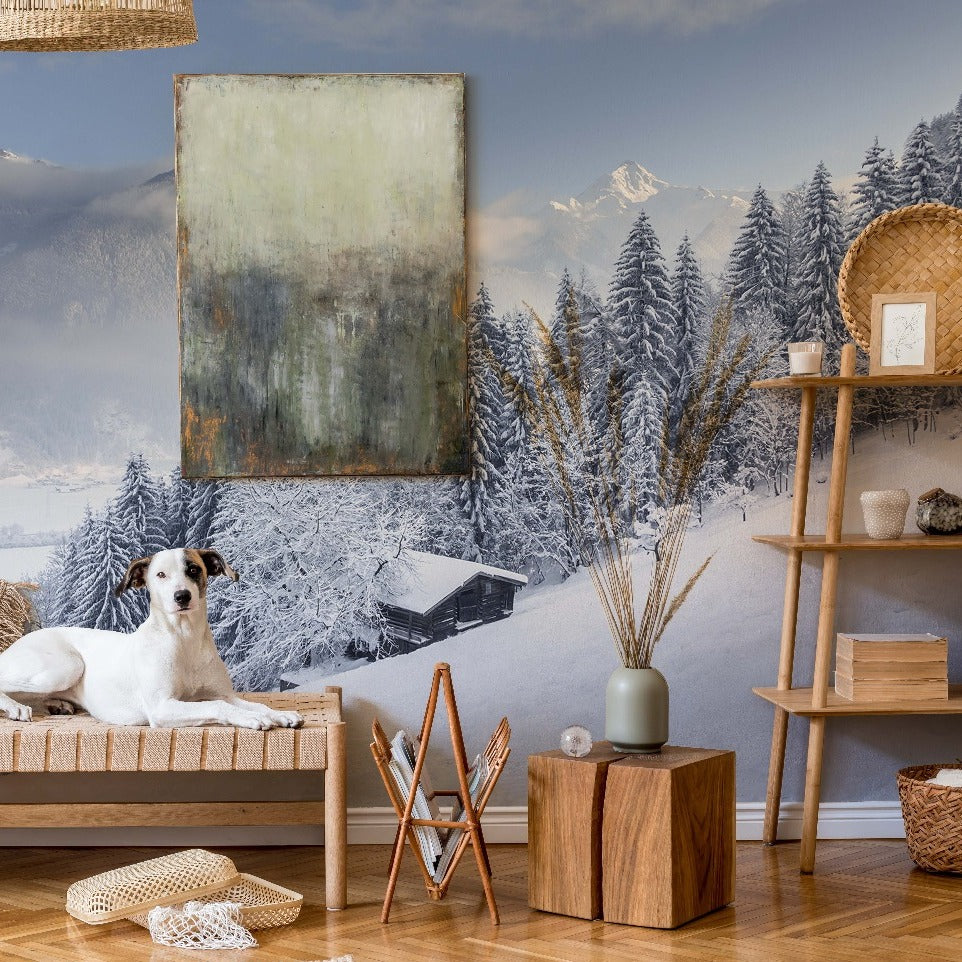 A cozy room with a Winter is Coming Wallpaper Mural by Decor2Go featuring snow-capped forest and trees, with a white dog laying on a bench, a large abstract painting, a shelf with decorations, and a wooden floor