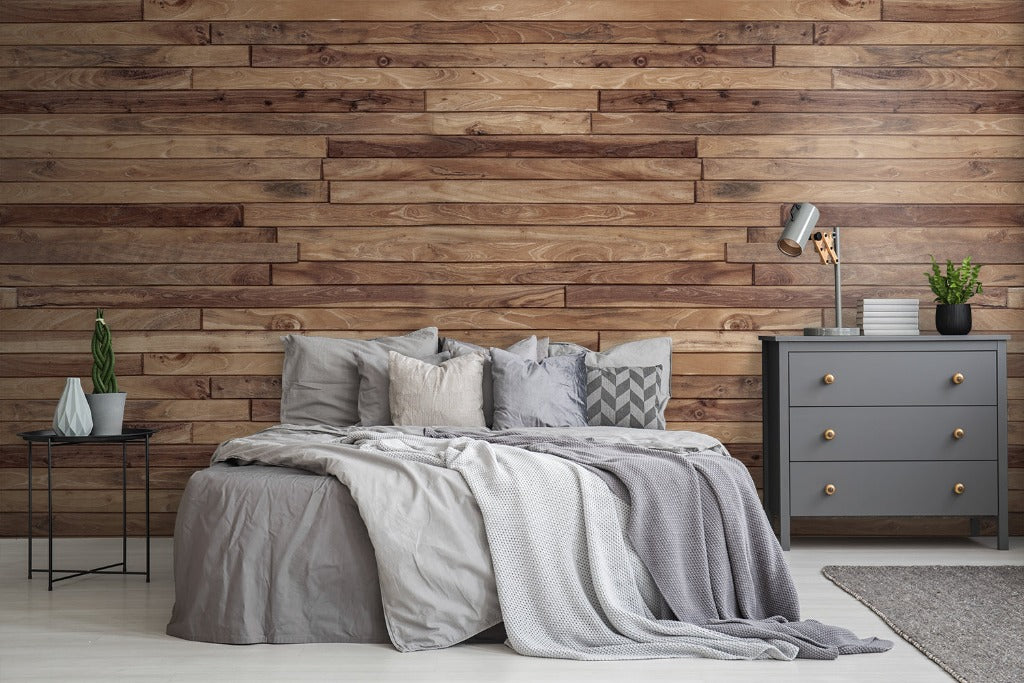 A cozy modern bedroom featuring a neatly made bed with gray linens, a Decor2Go Wallpaper Mural accent wall, a gray dresser, a small table lamp, and decorative plants.