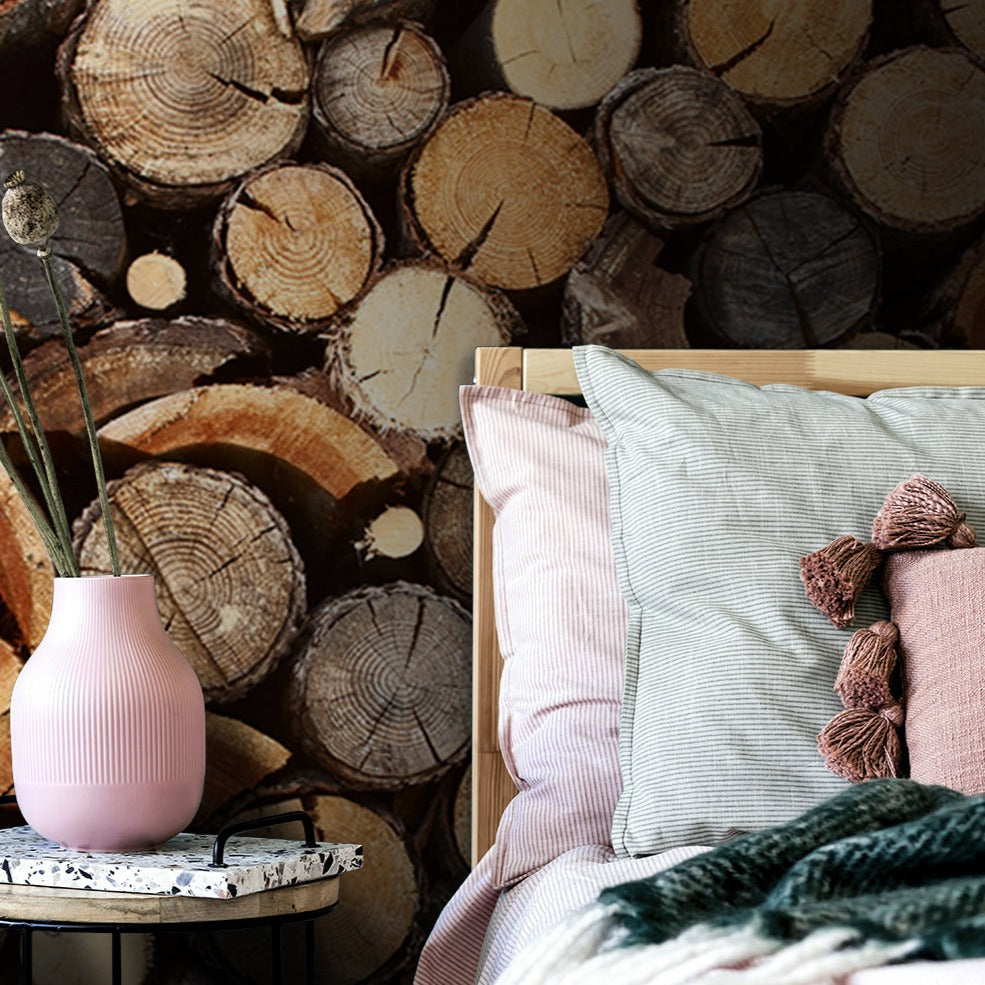 A cozy home interior featuring a stack of cut wood logs against a wall next to a stylish corner with a Decor2Go Wallpaper Mural on a small table, textured pillows, and a green knitted throw, all
