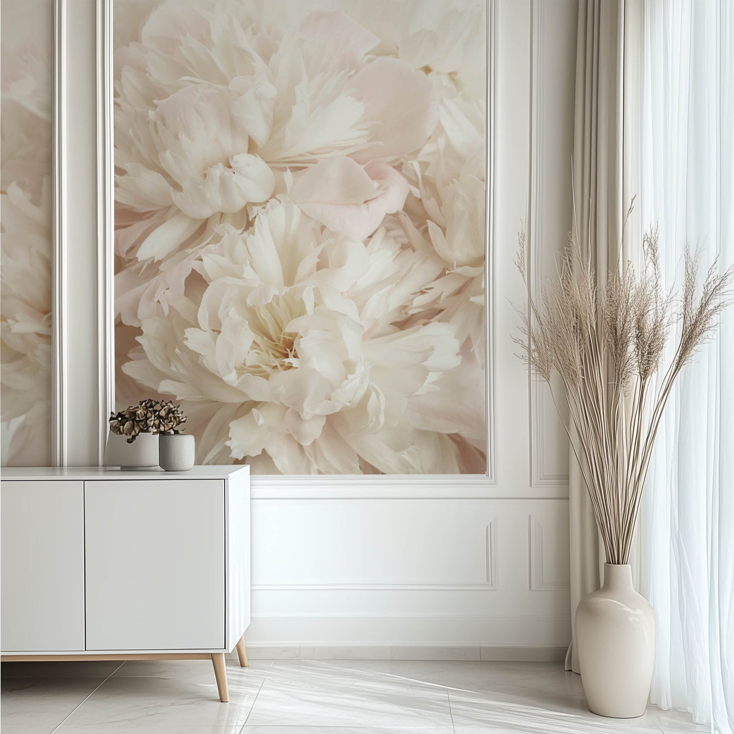 A minimalist living room showcases the Whispers of Peonies Wallpaper Mural by Decor2Go on the wall, complemented by a white sideboard with a small plant and a floor vase filled with tall pampas grass beside the sheer curtained window.