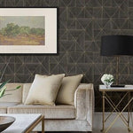 Enhancing a modern living room, a beige velvet sofa with two pillows is paired with a side table illuminated by a black and gold lamp. A small vase of white flowers adds elegance against York Wallcoverings' Black and Gold Cheverny Geometric Wood Wallpaper, accentuated by framed landscape artwork.