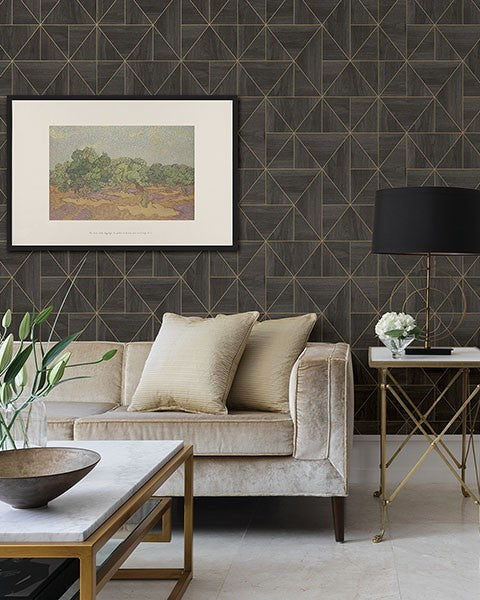 A modern living room with a beige velvet couch adorned with cushions in neutral tones. A framed landscape painting hangs on the Cheverny Geometric Wood Wallpaper (56 SqFt) by York Wallcoverings accent wall. The room also features a gold side table with a lamp and a small vase of flowers.