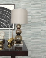 A modern table lamp with a beige shade and a base consisting of three stacked glass spheres sits on a dark wooden table. Next to the lamp are decorative items, including two candlesticks and a seashell. A large black and white abstract artwork hangs on the wall adorned with Lithos Geometric Marble Wallpaper (56 SqFt) by York Wallcoverings behind.