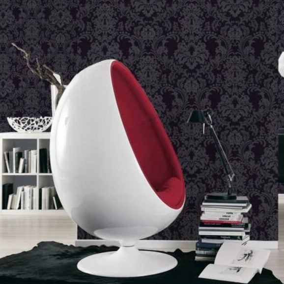 In a modern room design, an egg-shaped chair with a white exterior and red interior sits stylishly alongside a stack of books, a sleek black lamp, and a decorative vase on the bookshelf. The elegance is enhanced by Ontario's Black Baroque Black Wallpaper, which adds intricate texture to the walls, while the floor is elegantly unified with a black rug.