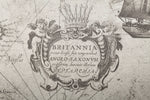 A detailed section of an antique map featuring a decorative emblem with the text "BRITANNIA prout divisa fuit temporibus ANGLO-SAXONUM, praefecturis ibernis illorum." The emblem is flanked by cherubs, reminiscent of Palitra's Voyage Wallpaper (114 SqFt) designs, and a ship is partially visible in the top right corner.