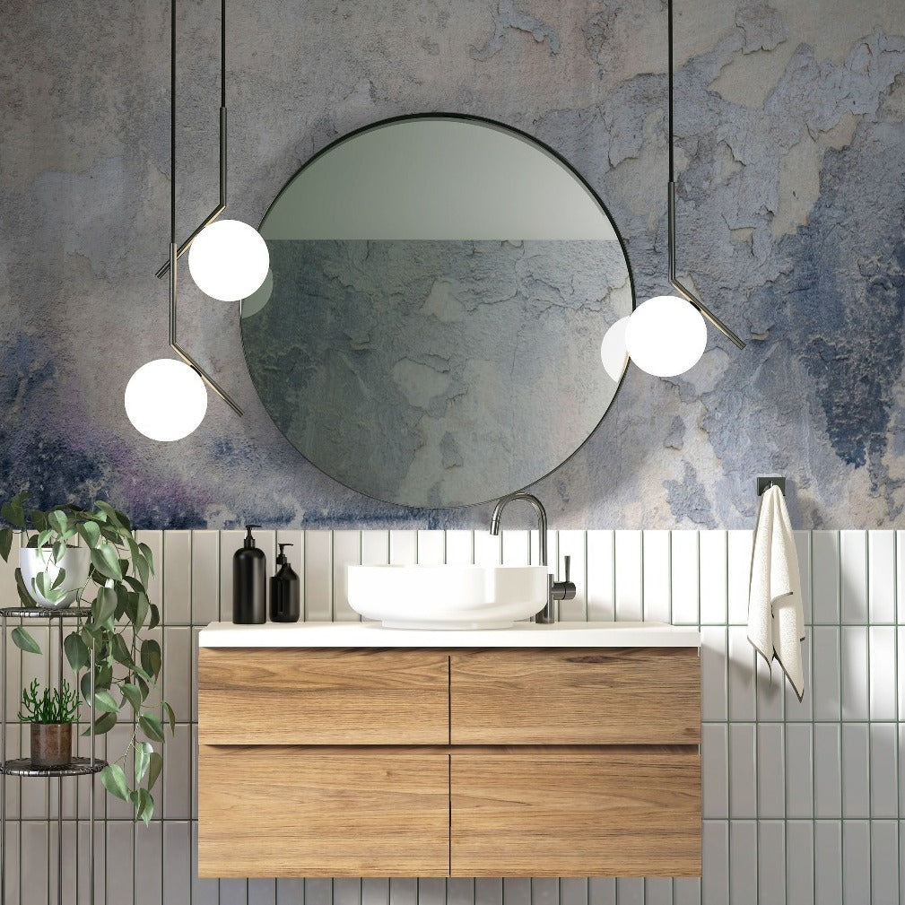 A modern bathroom vanity with a wooden cabinet and a white basin, flanked by pendant lights, set against a wall with tile and Decor2Go Wallpaper Mural. A round mirror and a plant add to the aesthetic.