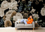 A woman in an orange sweater and jeans relaxing on a gray sofa, reading a book, against a dramatic backdrop of large Decor2Go Wallpaper Mural featuring peonies and blue flowers.