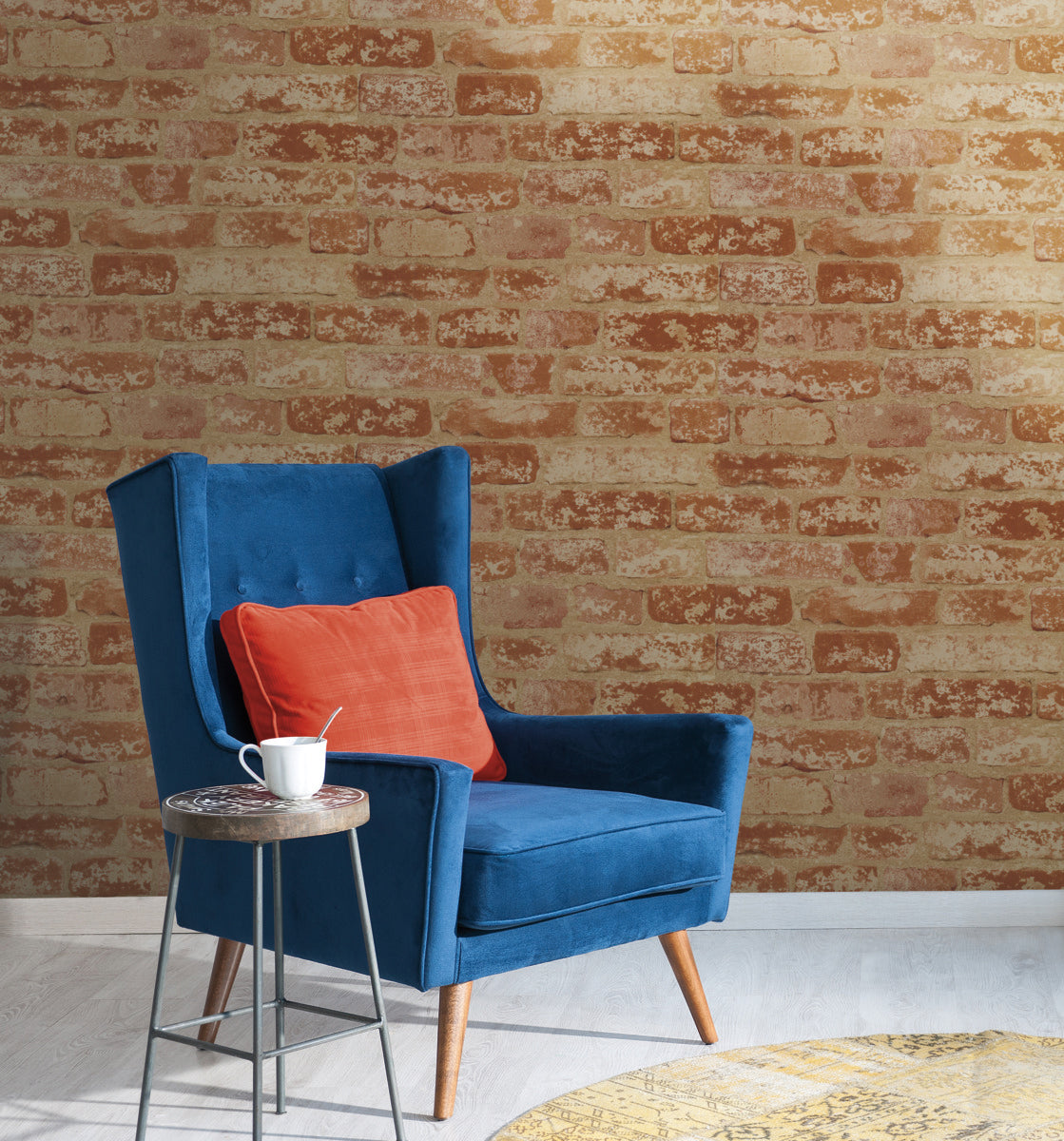 A cozy blue armchair with wooden legs sits against a wall adorned with York Wallcoverings' Urban Chic Up The Wall Wallpaper (56 SqFt). An orange cushion is placed on the chair. A small round side table beside the chair holds a white cup and saucer. The floor is light-colored, with a circular yellow rug partially visible.