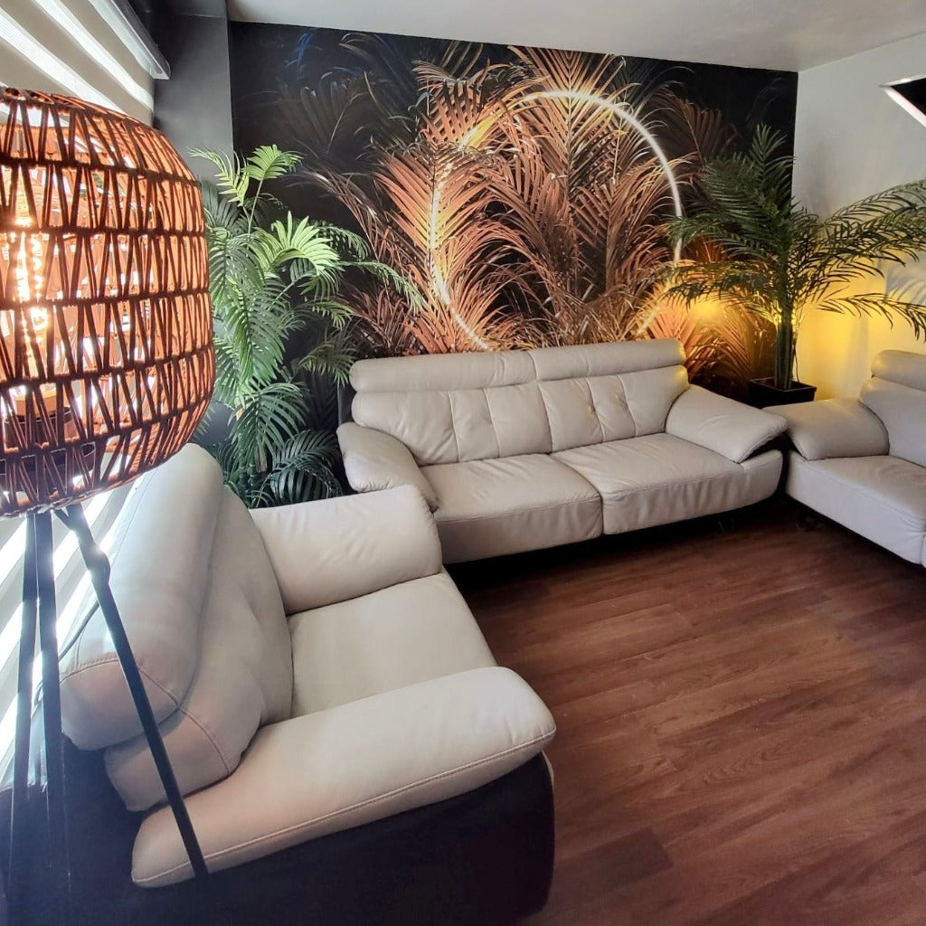 A stylish living room with a large "Big Green Leaves with Ring Light" wallpaper mural from Decor2Go, a beige sectional sofa, wooden floor, and a tall, woven floor lamp providing warm light.