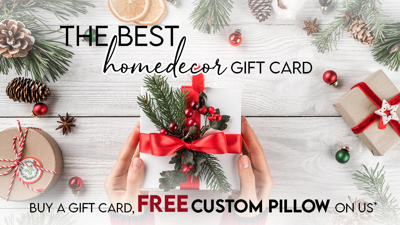 A person holds a gift box adorned with red ribbon and evergreen sprigs over a wooden table decorated with pine cones, evergreen branches, and ornaments. Text reads, "The Best Home Decor Gift Card from Decor2Go Winnipeg. Buy a $100 gift card, get a free custom pillow on us*.