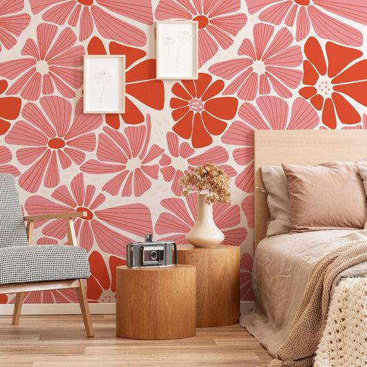 A cozy bedroom corner featuring a bed with pink linens, a Decor2Go Wallpaper Mural with a groovy daisy flower pattern, a wooden side table with a vintage camera, and a chair with checkered upholstery.