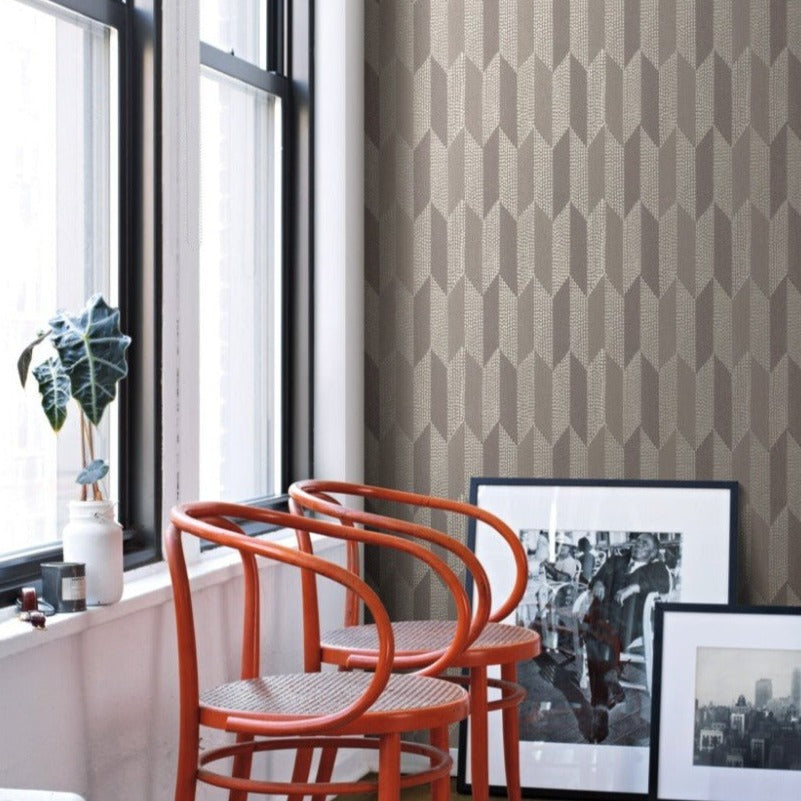 A cozy corner showcases two red chairs with woven seats, a potted plant on the windowsill, and framed black-and-white photos leaning against York Wallcoverings' Cosmopolitan Wallpaper in Dark Oyster, which adds a subtle raised texture. Natural light streams in through the large windows.