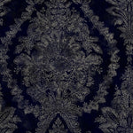 Charleston Damask Elegant Wallpaper by York Wallcoverings transforms ornate dark blue fabric into a masterpiece, featuring silver floral and leafy designs. It showcases detailed flower motifs and curved foliage for a sophisticated, symmetrical look. Available in 60 SqFt rolls.