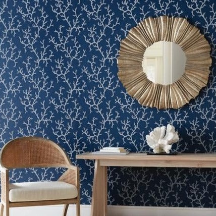 A cozy room featuring a wooden chair with a white cushion, a wooden desk adorned with a shell and a blue book, and a round mirror with a gold sunburst frame. The wall behind the desk and chair is covered with Coral Island (60 SqFt) Wallpaper by York Wallcoverings showcasing blue hues and white branch patterns.