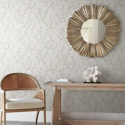 A minimalist room features a light wooden chair with a woven backrest and cushioned seat, a wooden console table with a small white decorative object, and a round mirror with a textured, wavy gold frame on the wall behind, adorned with subtle Coral Island Wallpaper (60 SqFt) by York Wallcoverings.