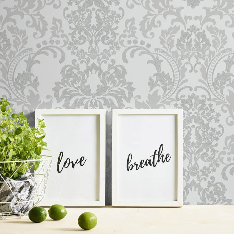 Two framed prints with the words "love" and "breathe" lean against Ontario's White Baroque White and Light Grey Wallpaper. A small potted plant and a few limes rest on the textured surface of durable material in front of the frames.