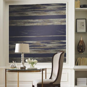 Home Office - A stylish features a modern painting with horizontal blue and white streaks, centered on a beige wall. The room includes a wooden desk with a white lamp and a potted plant, and a beige upholstered chair. Shelves with books and decor items are to the right, all set against Dry Brush Wallpaper (60 SqFt) by York Wallcoverings