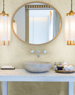 A modern bathroom vanity features a round mirror with wooden trim, a stone sink, and two modern pendant lights on either side. The countertop holds toiletries, a soap dispenser, a decorative tray with rolled towels, and a soap dish. The wall is adorned with York Wallcoverings' Geodes Wallpaper (60 Sq Ft).