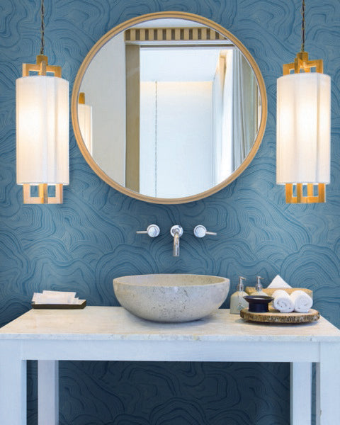 Bathroom - A modern features a round mirror above a white marble sink. Two cylindrical pendant lights with white shades and wooden accents flank the mirror. The walls boast a blue, textured pattern resembling Geodes Layers Wallpaper (60 SqFt) by York Wallcoverings. Accessories include towels, soap, and a small bowl on the counter