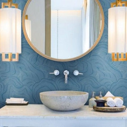 A modern bathroom features a round mirror with a wooden frame, mounted on York Wallcoverings' Geodes Wallpaper (60 Sq Ft) with a blue textured pattern. Below the mirror is a stone vessel sink with a wall-mounted faucet. Two contemporary pendant lights hang on either side. Various toiletries are arranged on the countertop.