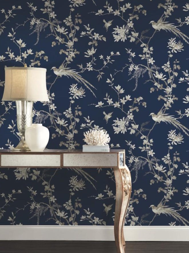 A decorative table with a metallic finish displays a lamp featuring a white shade alongside a white coral ornament. The backdrop showcases York Wallcoverings' Blue Bird And Blossom Chinoiserie Wallpaper, which adds an elegant touch with its dark blue hue and intricate white floral and bird designs.