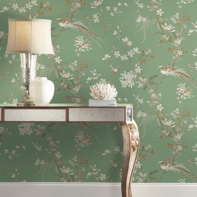 A decorative table with a mirrored, metallic finish is positioned against York Wallcoverings' Green Bird and Blossom Chinoiserie Wallpaper, featuring intricate bird and branch patterns. The elegant scene showcases a lamp, a white vase, and a coral ornament atop the table.