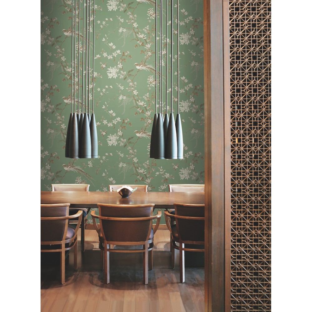 A dining area with a wooden table surrounded by chairs, set against vibrant Bird And Blossom Chinoiserie Wallpaper (60 SqFt) by York Wallcoverings. Three black pendant lights with a metallic finish hang above the table. A decorative wooden lattice screen is visible on the right side of the image.