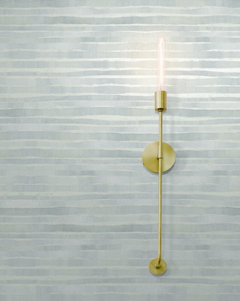 A minimalistic wall sconce with a tall, slender brass design and an exposed bulb is mounted on a pale blue and white striped Dreamscapes Stripes (60 SqFt) by York Wallcoverings. The light cast by the bulb gently illuminates the surrounding area.