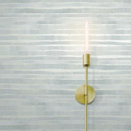 A wall-mounted brass light fixture against a gray, horizontally striped **Dreamscapes Wallpaper (60 SqFt) by York Wallcoverings**. The fixture features a single exposed, vertically oriented, elongated bulb, emitting a soft, warm glow that adds a touch of metallic shimmer to the scene.