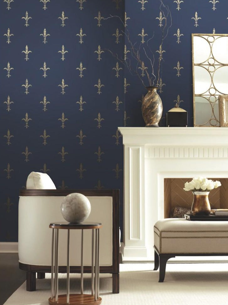 An elegant living room featuring a dark blue Fleur De Lis (60 SqFt) vinyl-coated wallpaper with a golden floral pattern, a white fireplace, and stylish furnishings including a mirror, a chair, and decorative vases by York Wallcoverings.