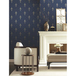 A stylish living room with dark blue Fleur De Lis Wallpaper (60 SqFt) by York Wallcoverings featuring fleur-de-lis patterns. A white fireplace adorns the wall, along with a mirror and decorative vases. A white armchair, a side table with a spherical decor, and a beige cushioned bench complete the scene.