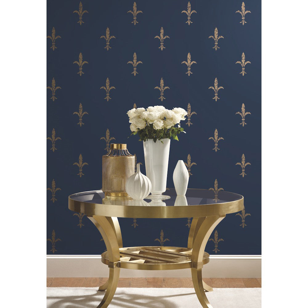 A modern room with dark blue, York Wallcoverings Fleur De Lis wallpaper adorned with golden floral patterns, featuring a shiny gold circular table holding a white vase with white flowers and decorative items.