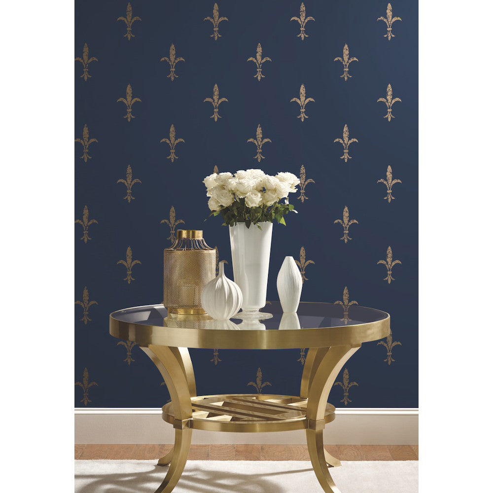 A stylish round table with a gold finish stands against a dark blue wall adorned with golden fleur-de-lis patterns on Fleur De Lis Wallpaper (60 SqFt) from York Wallcoverings. On the table are a white vase filled with white flowers and decorative items, including a gold container and a white ceramic onion-shaped object.
