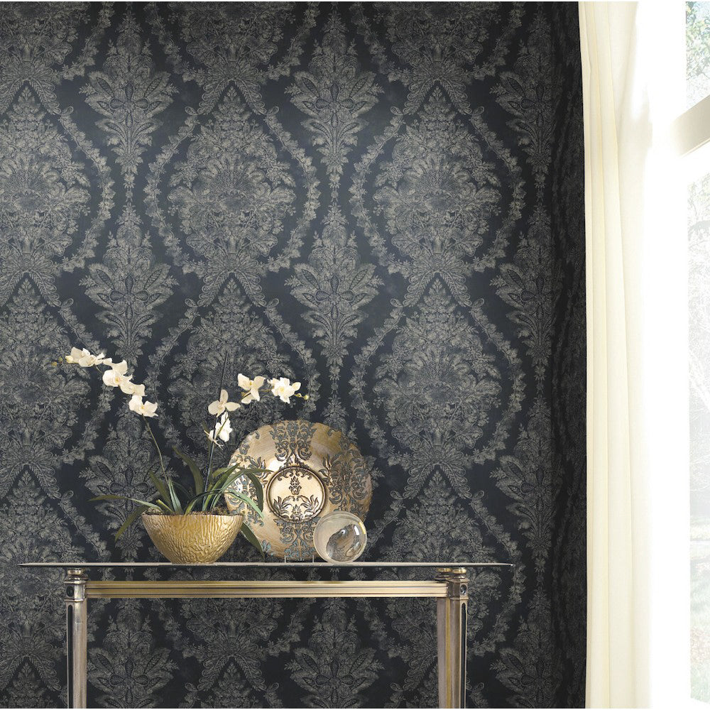 A decorative accent table stands against a wall adorned with Classic Charleston Damask Wallpaper in Blue (60 SqFt) by York Wallcoverings. The table displays a potted plant with white orchids, a golden bowl, a decorative plate, and a clear orb. Natural light filters in through a window, highlighting the intricate pearlescent floral motifs.